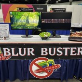 Trade show booth for Blur Busters with two computer monitors on a table, one showing a forest wallpaper and the other a dark screen, plus a QR sign on the left.