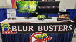 Trade show booth for Blur Busters with two computer monitors on a table, one showing a forest wallpaper and the other a dark screen, plus a QR sign on the left.